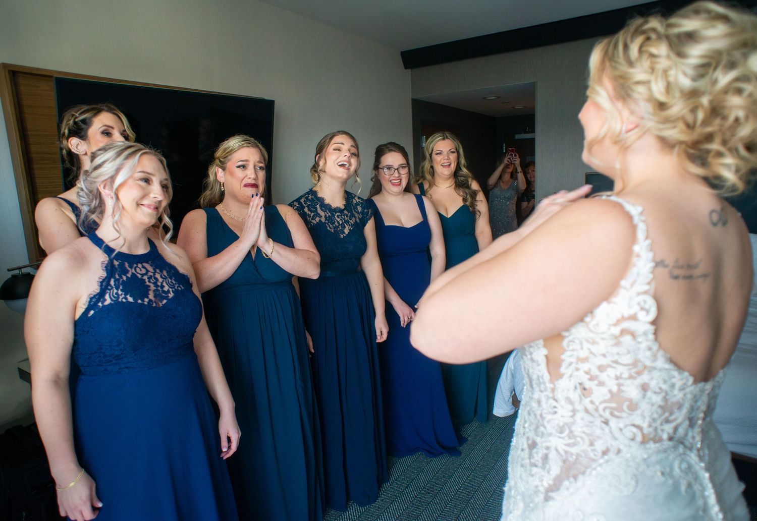 Bridesmaids in navy blue dresses react emotionally to seeing the bride in her wedding gown.