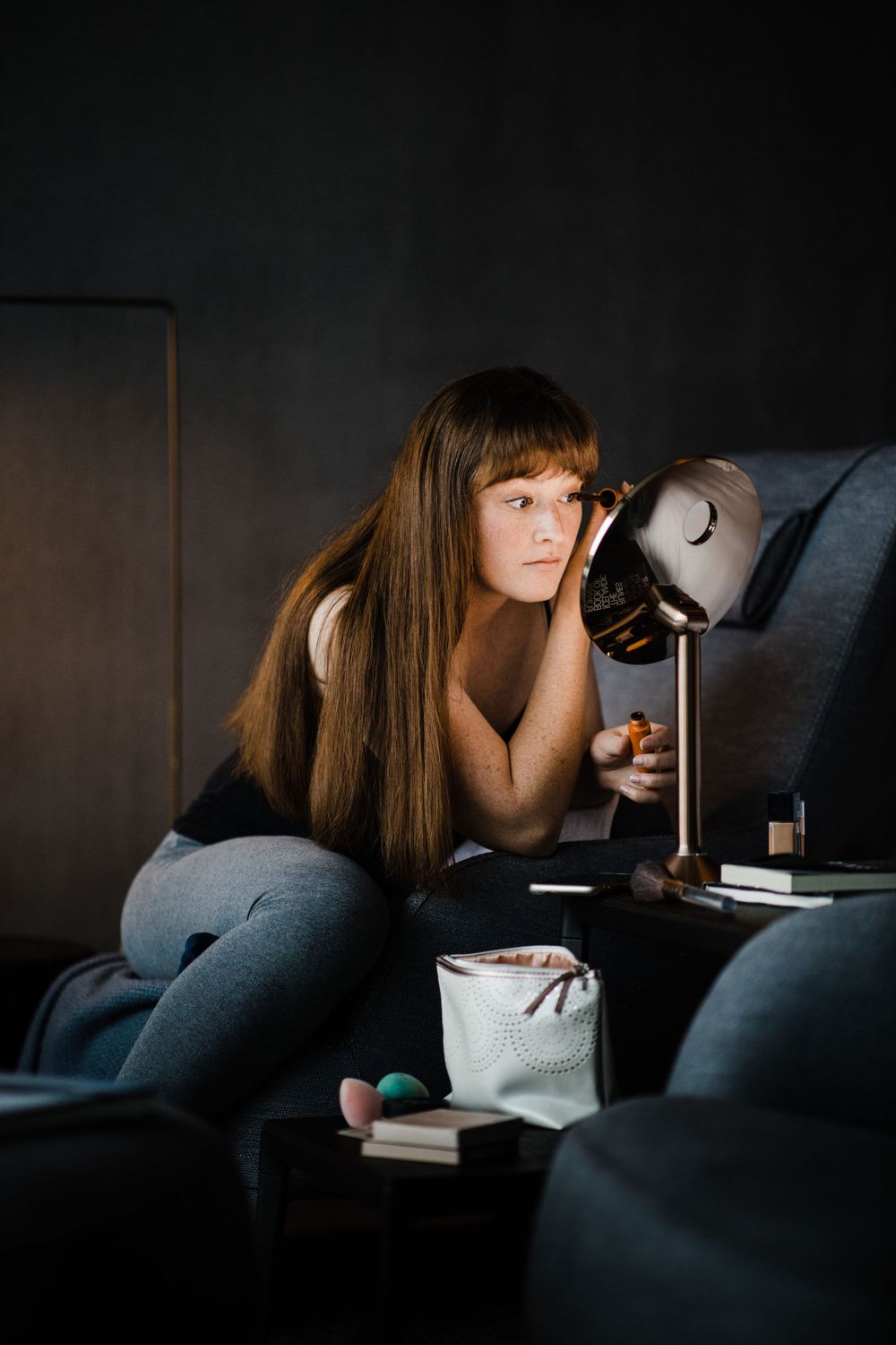 A person lounges on a couch reading by lamplight in a dark room.