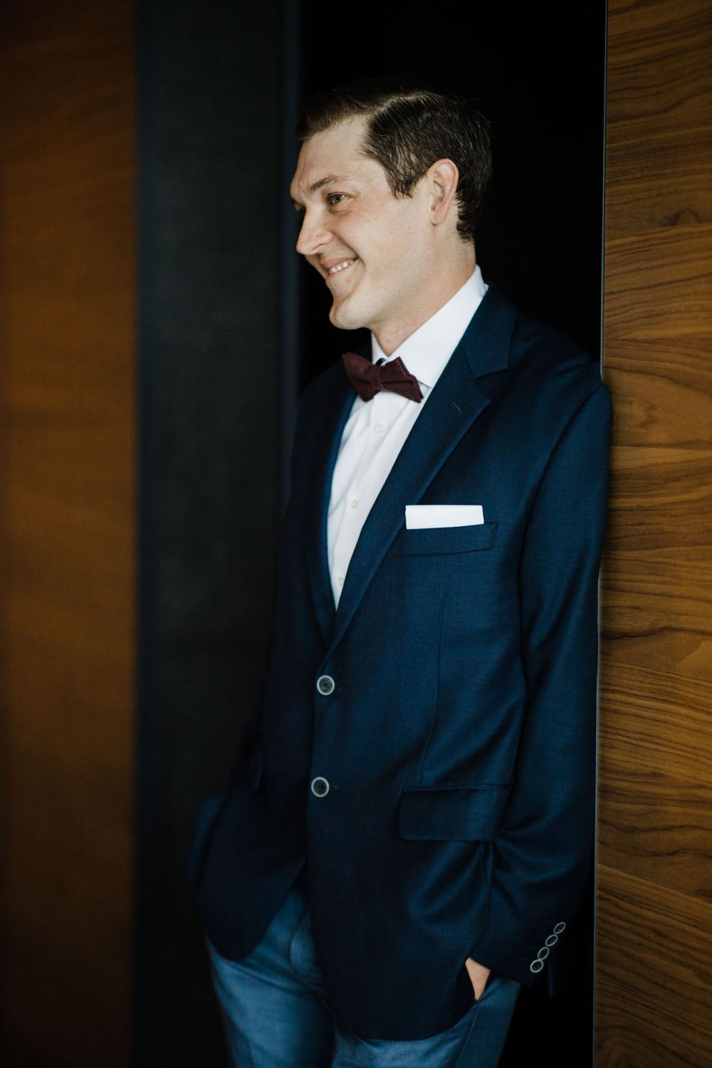 Man in formal navy suit and bowtie smiles while standing against wooden wall at formal event.