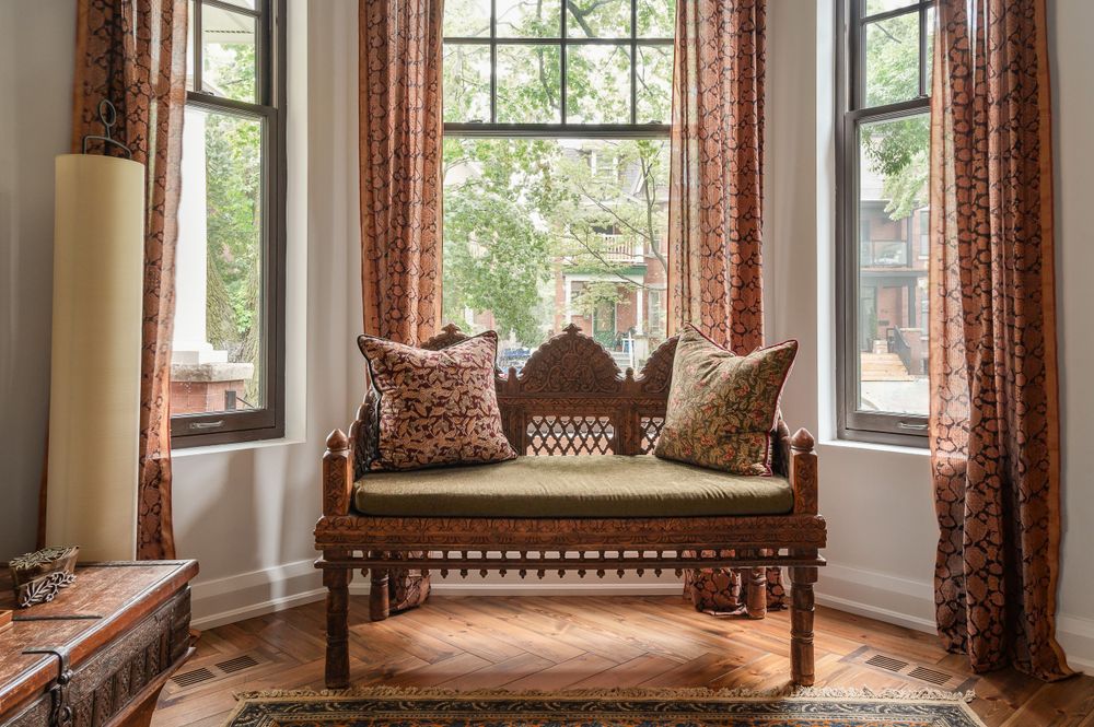 Elegant wooden bench with carved details in a bay window nook with patterned curtains.