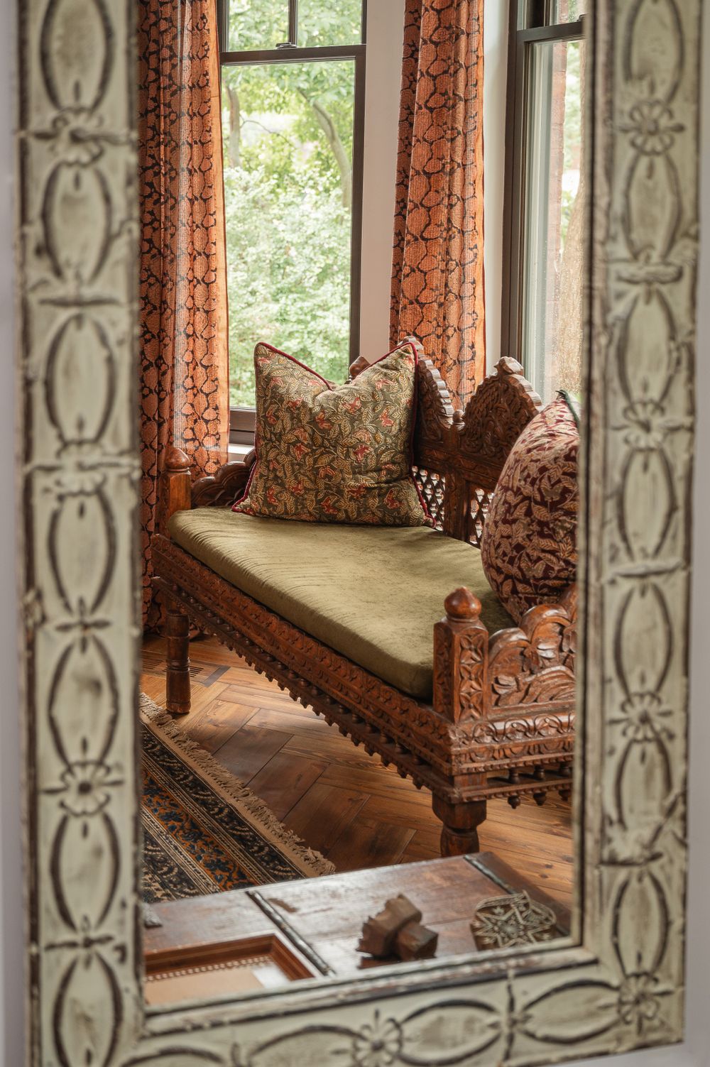 Ornate carved wooden bench with floral cushions seen through a decorative mirror frame.
