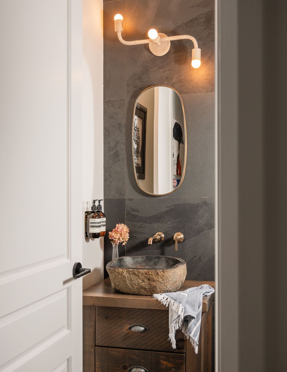 Modern bathroom with stone vessel sink, curved lighting fixture, and oval mirror on dark grey wall.