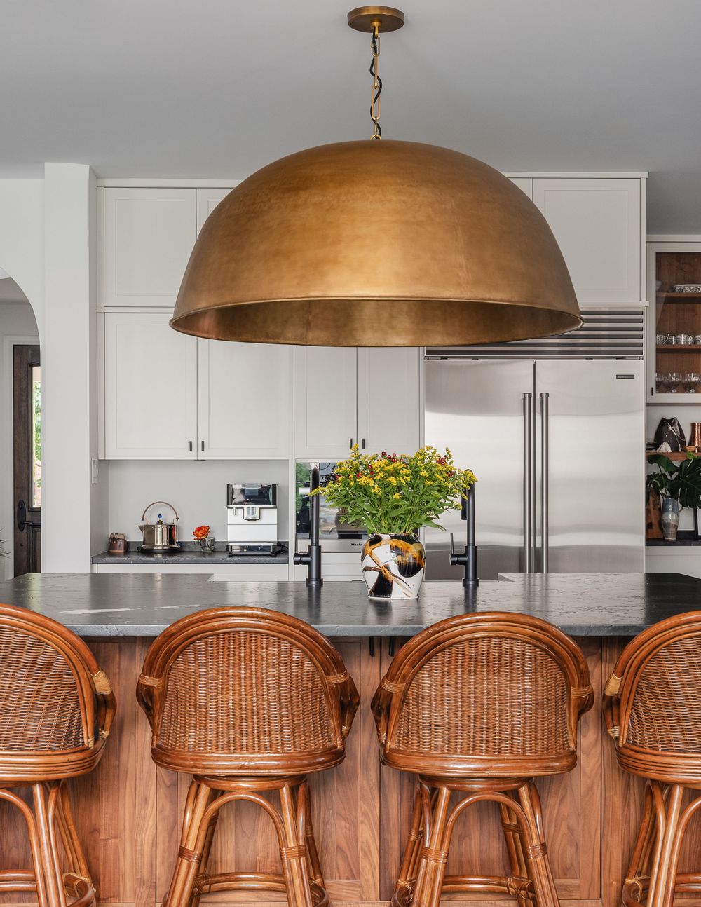 Modern kitchen with large brass dome pendant light over wood and rattan bar stools.