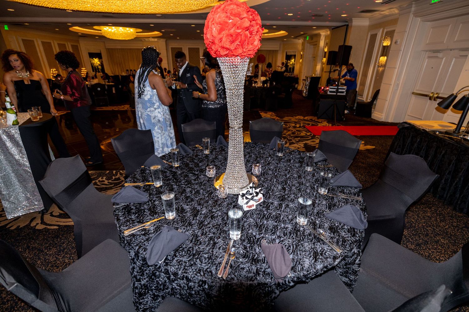 Formal event table with black linens and tall red floral centerpiece in an elegant ballroom setting.