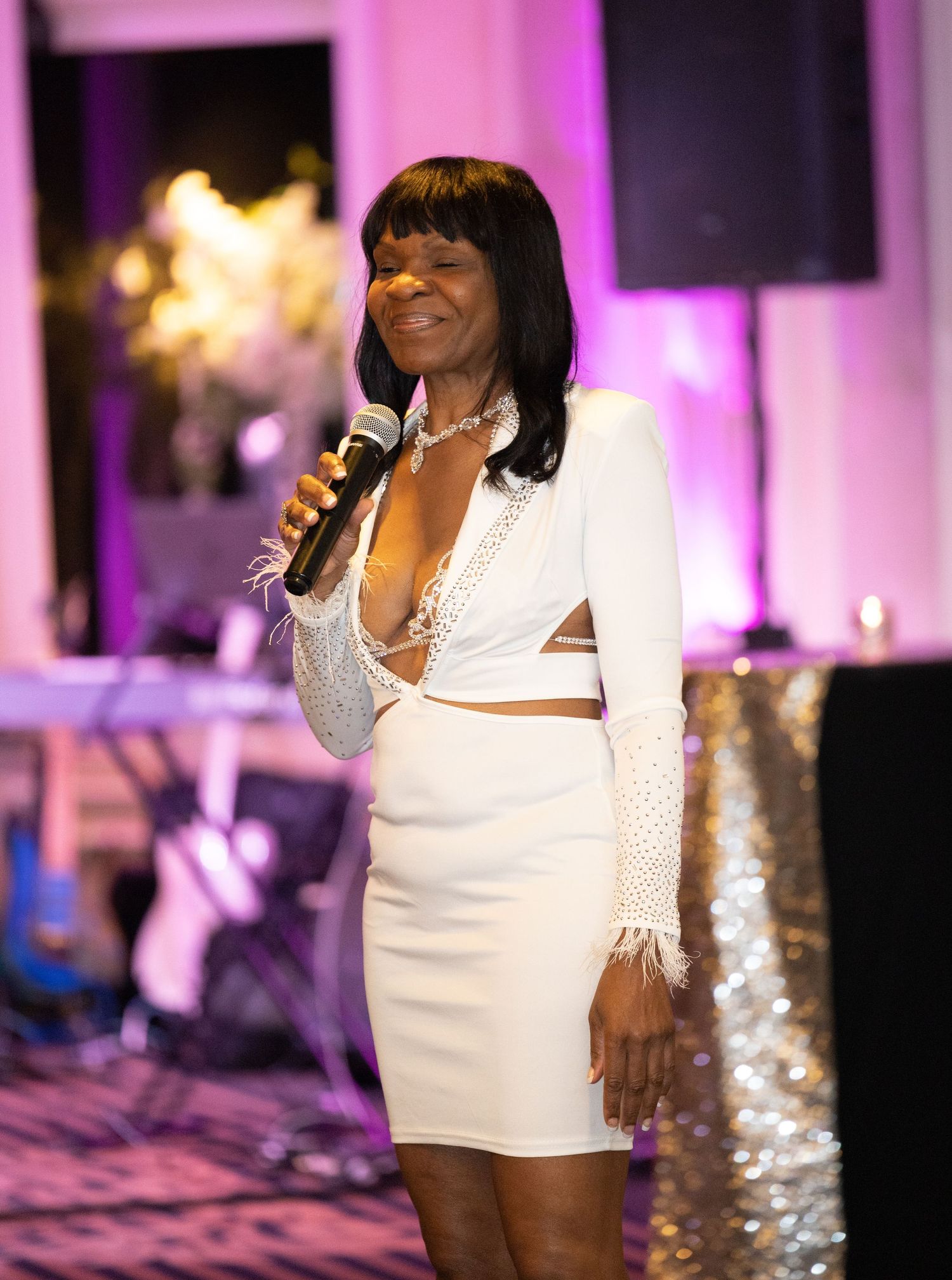 Performer in a white sequined outfit singing on stage with pink lighting effects.
