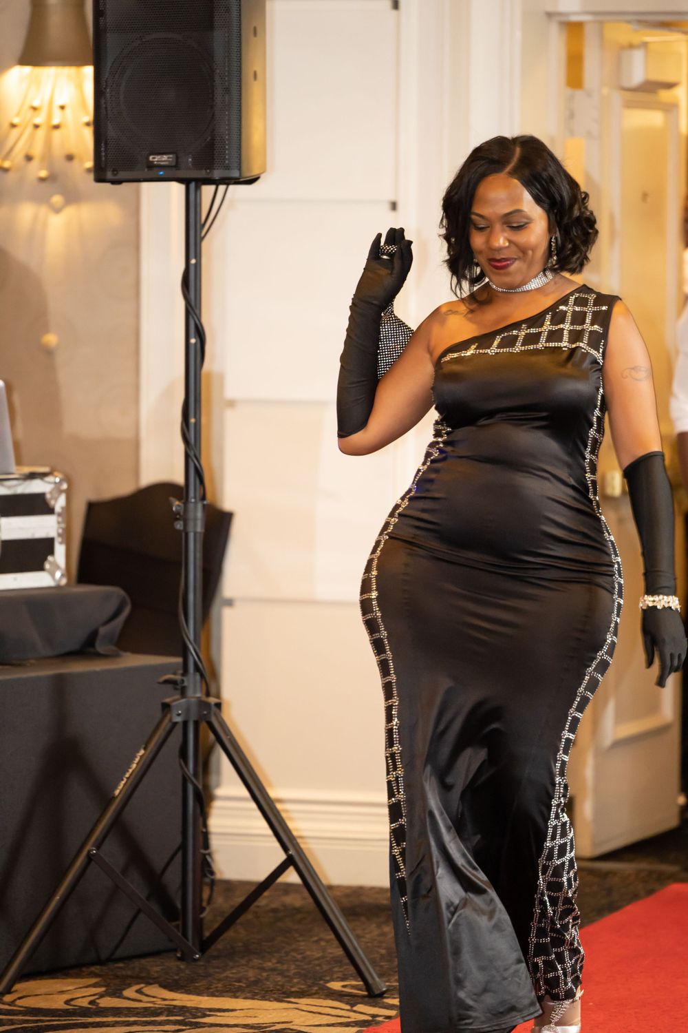 Woman in stunning black evening gown with metallic geometric pattern stands next to speaker at event.