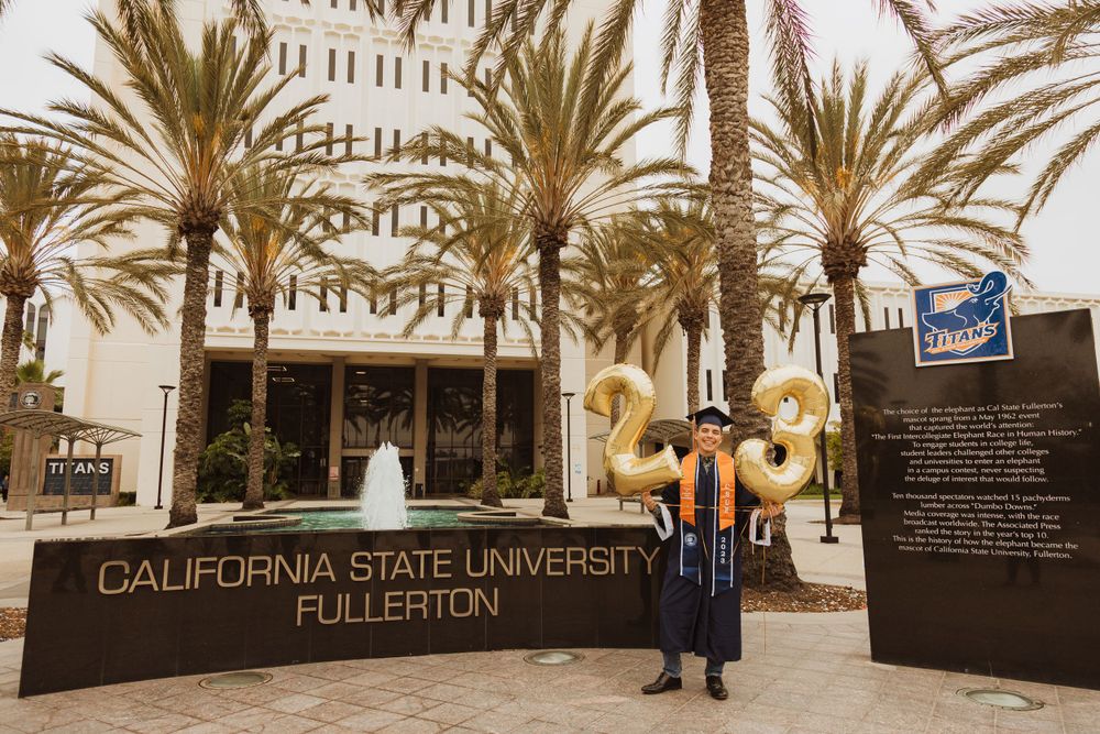 A graduate stands near the Cal State Fullerton entrance sign surrounded by palm trees.