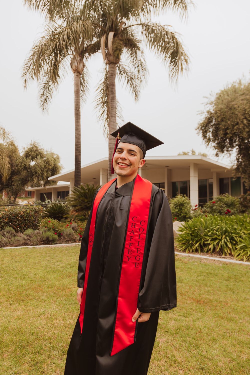 Graduate in black gown and red stole poses on lawn with palm tree and building in background.