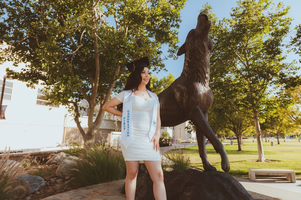 Graduate in white dress stands by rearing bronze horse statue surrounded by trees.