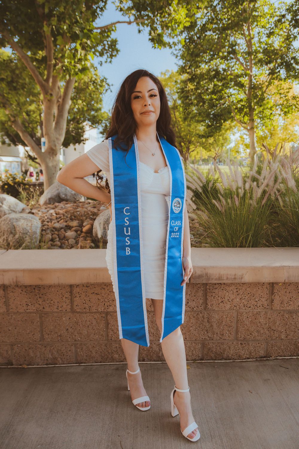 Graduate wearing blue stole and white dress posing against stone wall with greenery in background.