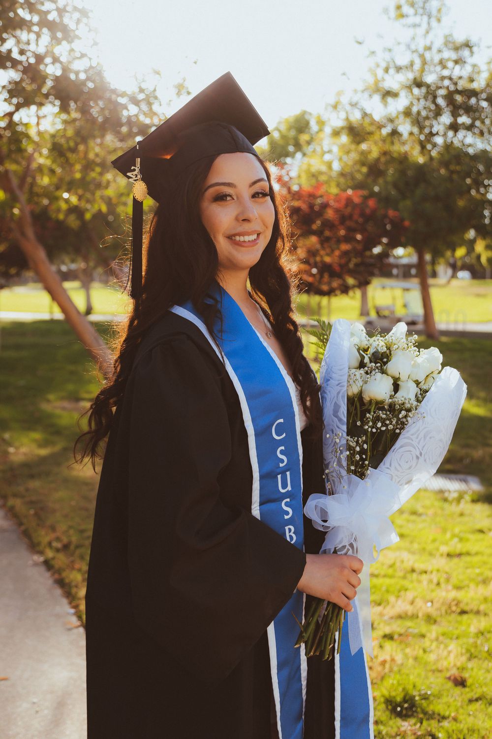 Graduate in cap and gown holding white roses while posing in sunny campus garden.