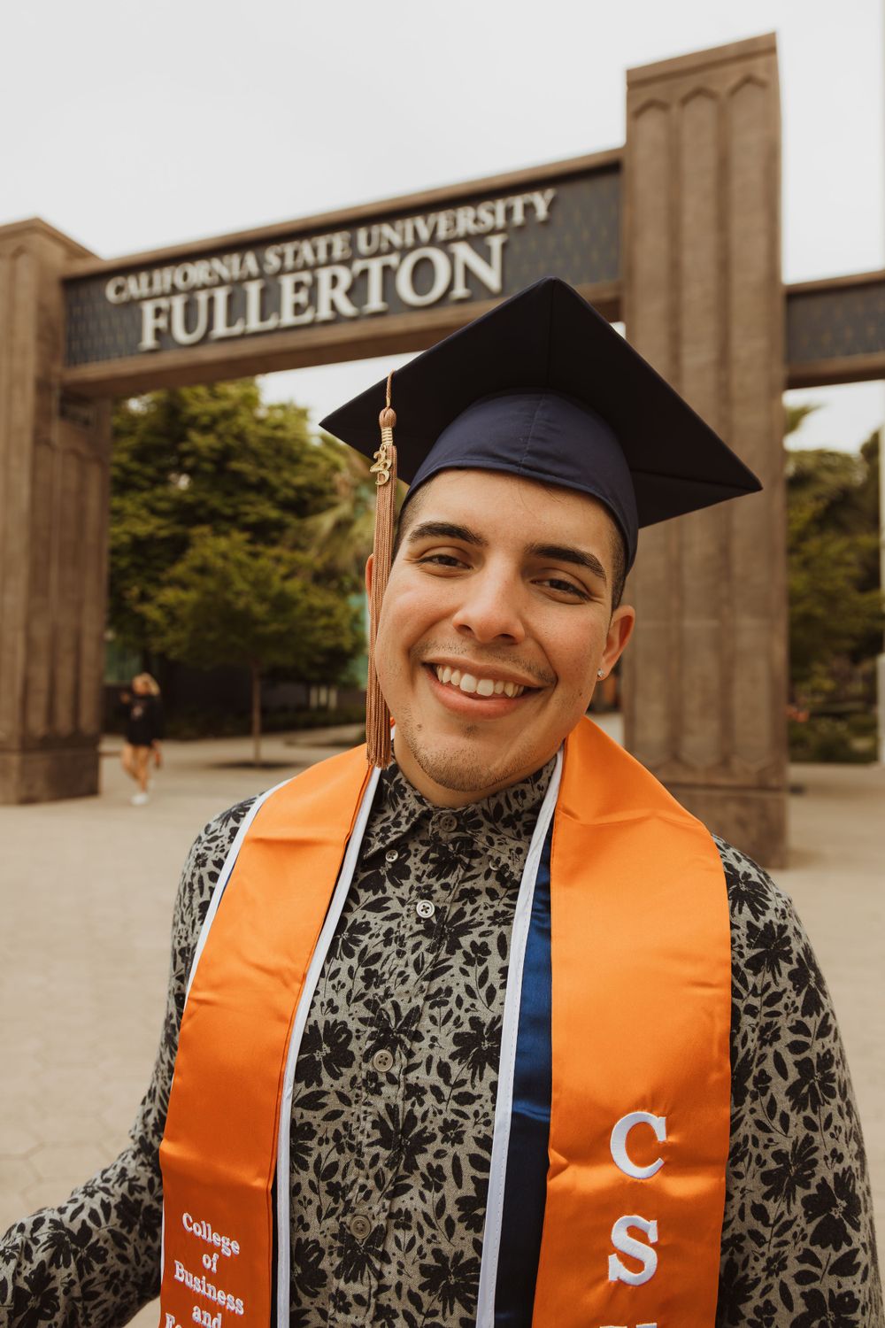 Graduate in cap and gown smiling in front of California State University Fullerton entrance sign.