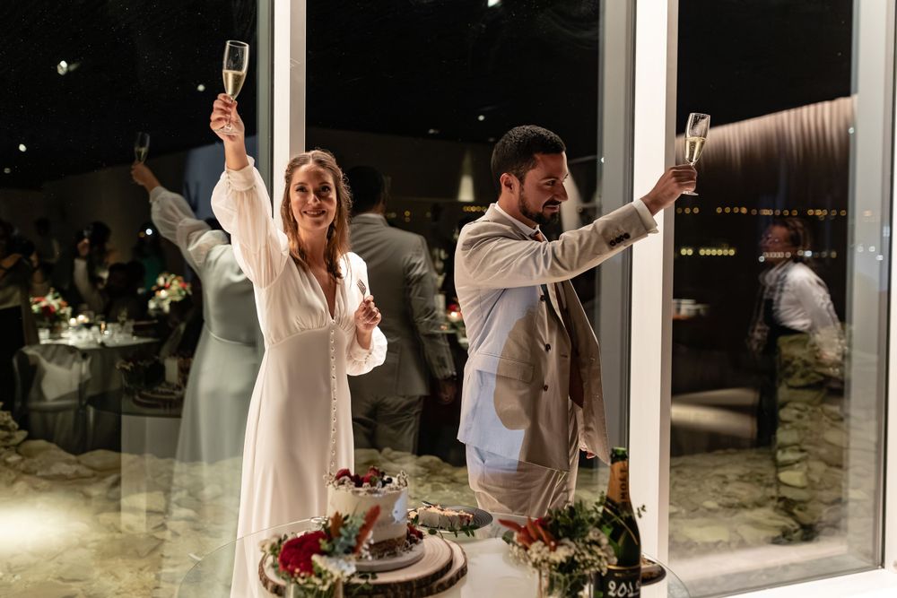 A couple raises their champagne glasses in celebration at their wedding reception by a glass window at night at Forte da Crismina, Cascais