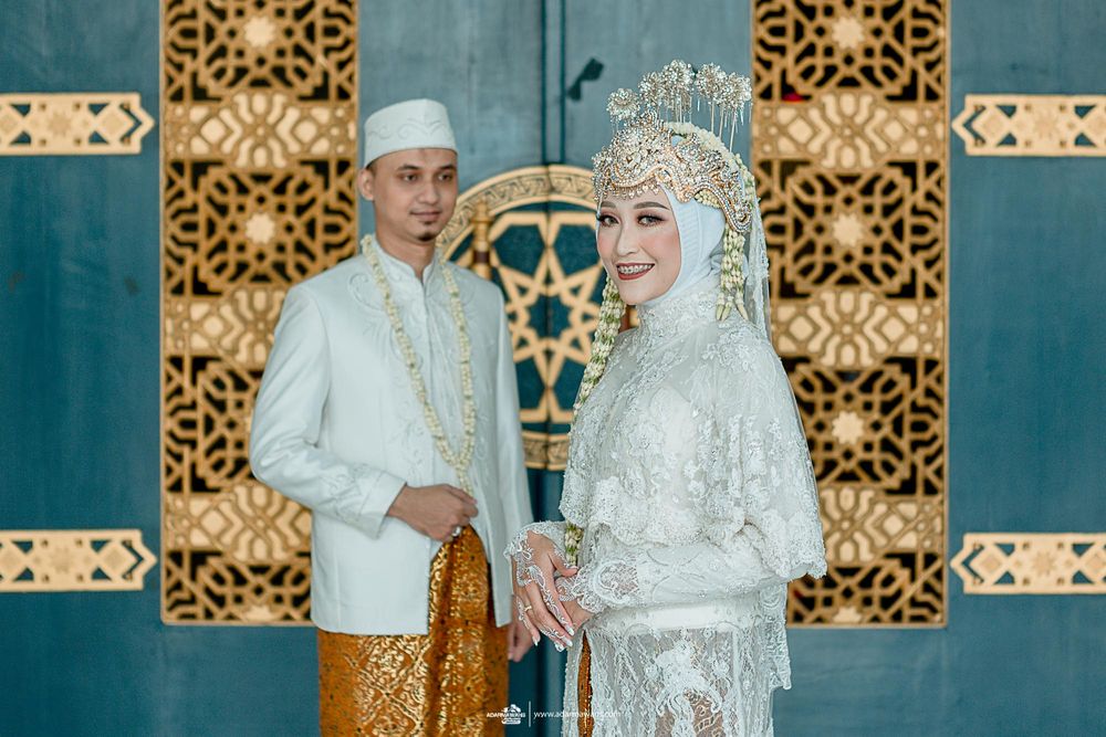 Traditional Indonesian wedding couple in white and gold attire standing against ornate patterned background.