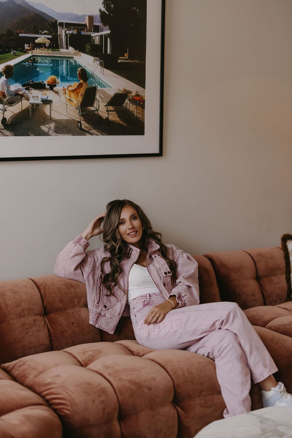 Relaxed indoor portrait on brown couch wearing pink denim outfit with pool artwork on wall.