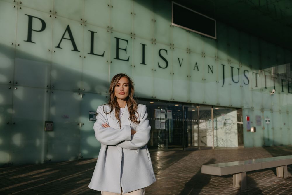 Fashion model in white coat poses against Paleis van Justitie building exterior.