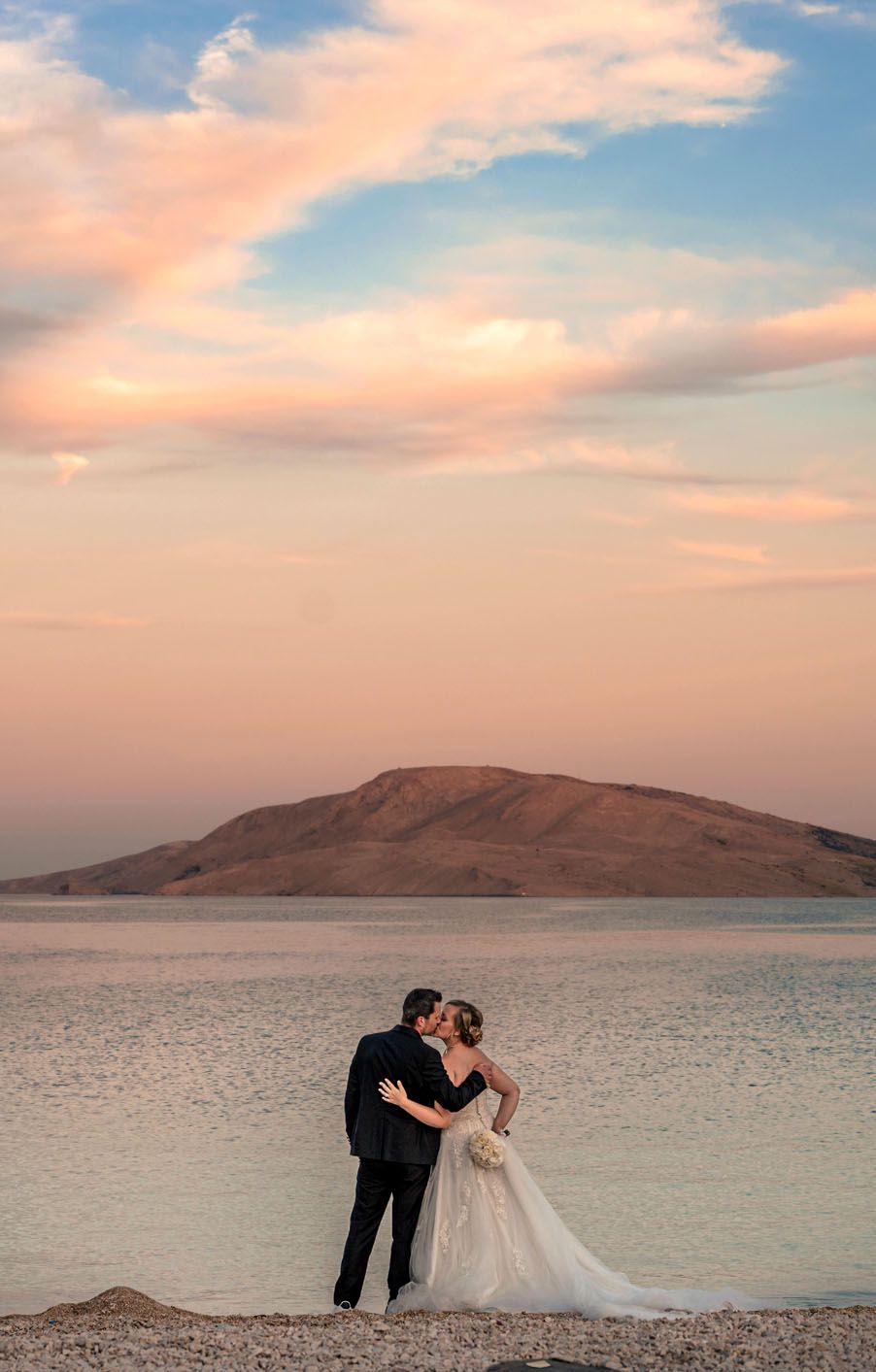 A couple poses against a scenic desert backdrop with mountains and water during golden hour.