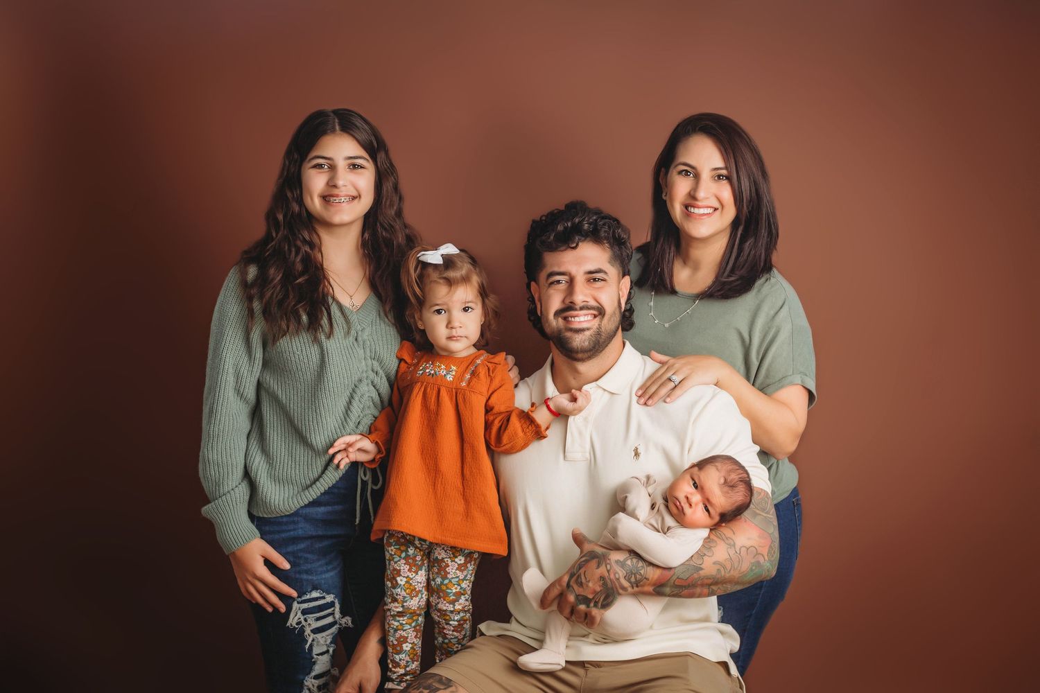 A studio family portrait with parents and children in casual attire against a warm brown background.