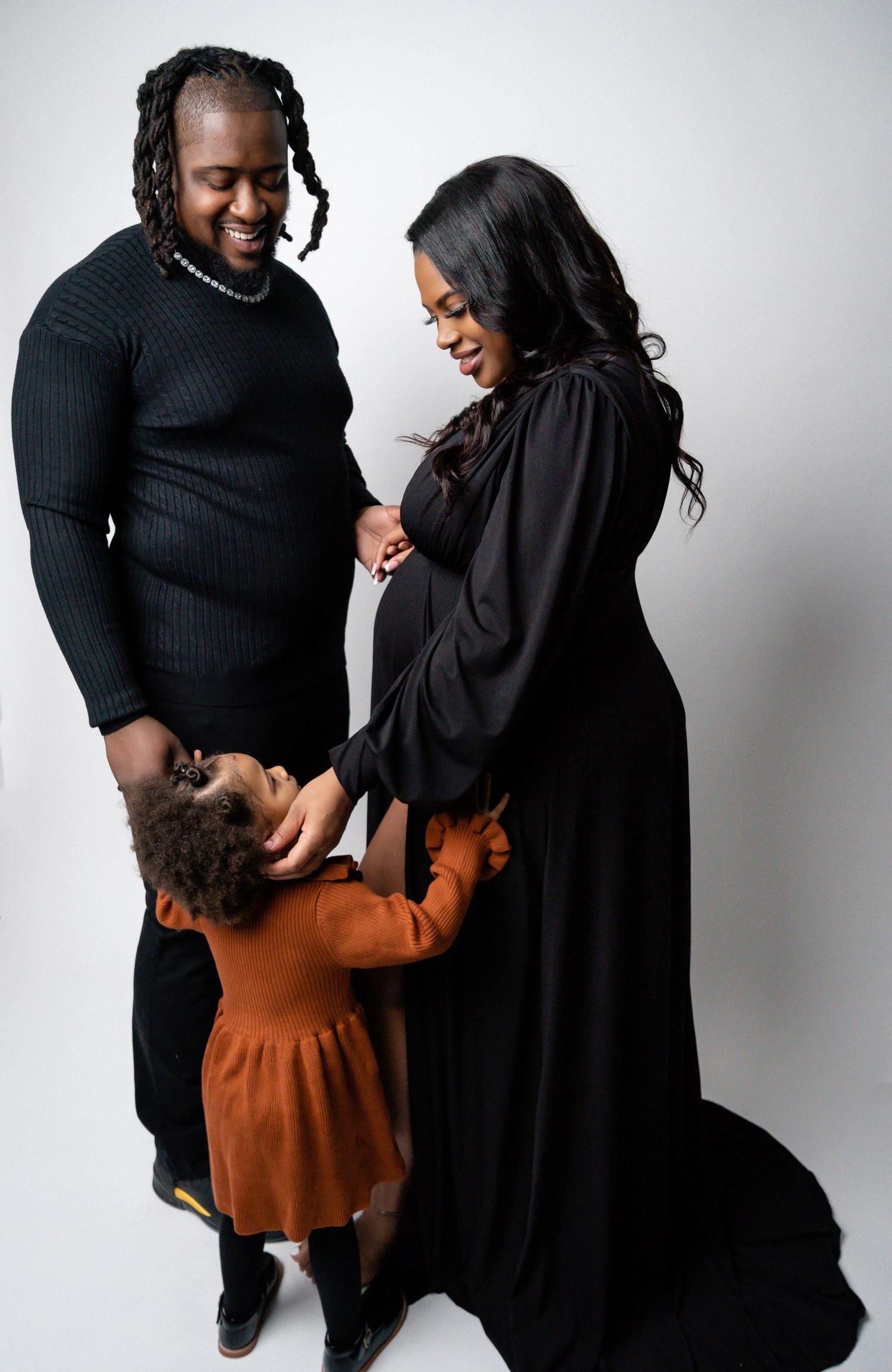 Family maternity photoshoot with couple in black and brown outfits against white backdrop.