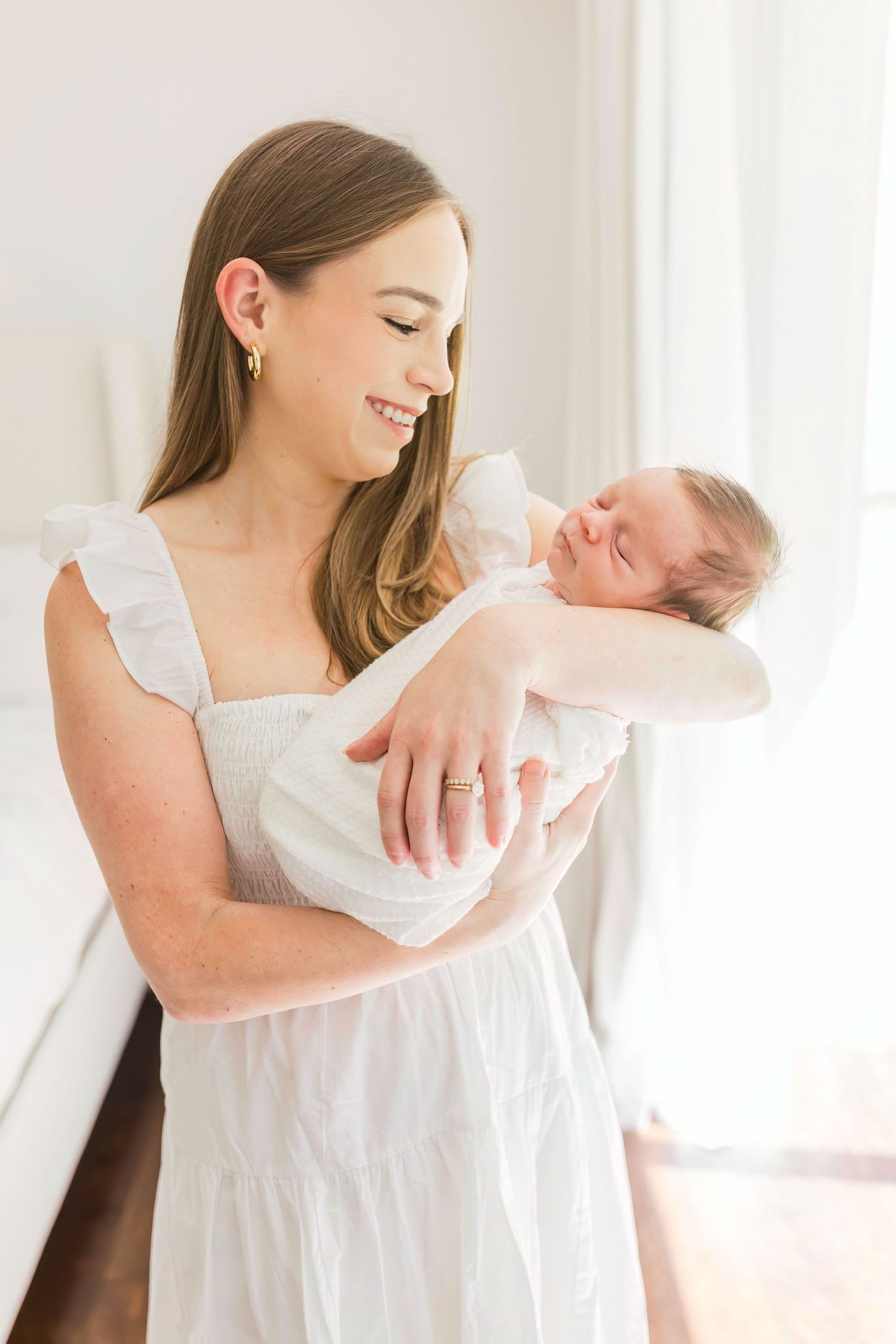 A mother in white dress smiles while cradling her sleeping newborn by a bright window.