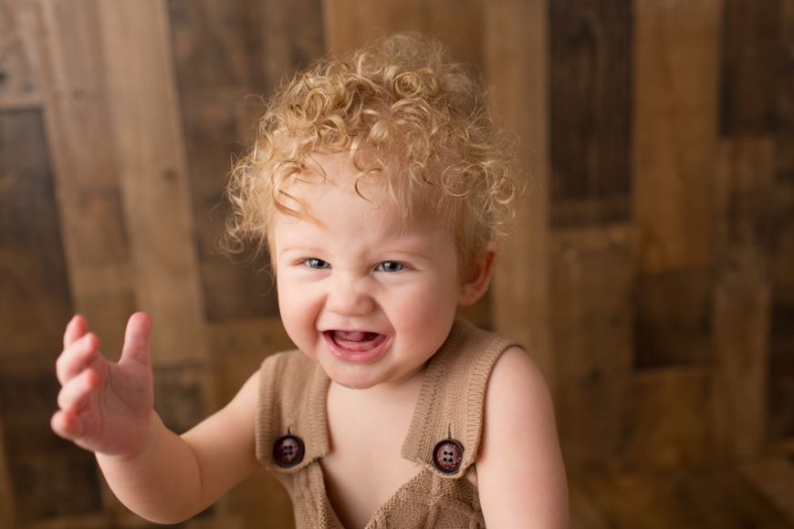 Close-up portrait of a toddler with curly hair wearing brown overalls against wooden backdrop.