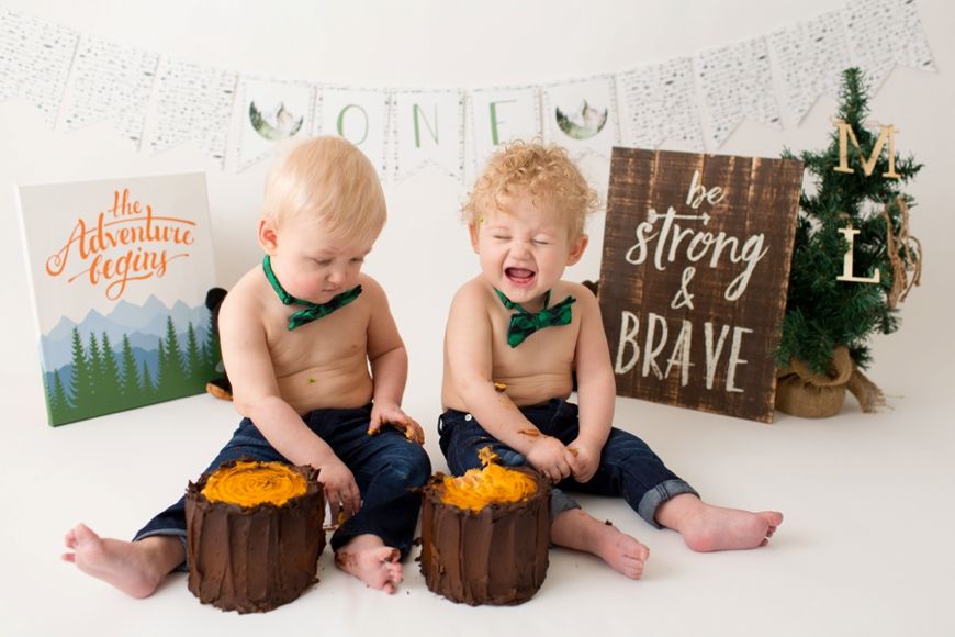 Two toddlers sitting by decorated tree stumps with orange cakes and woodland themed wall art.