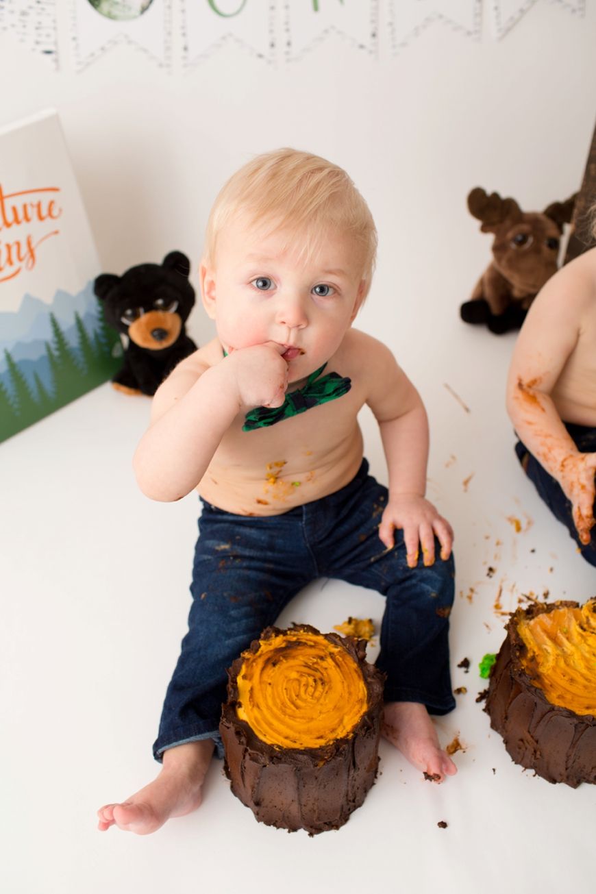 Toddler in jeans sitting on floor with orange frosted cake and woodland themed decorations.