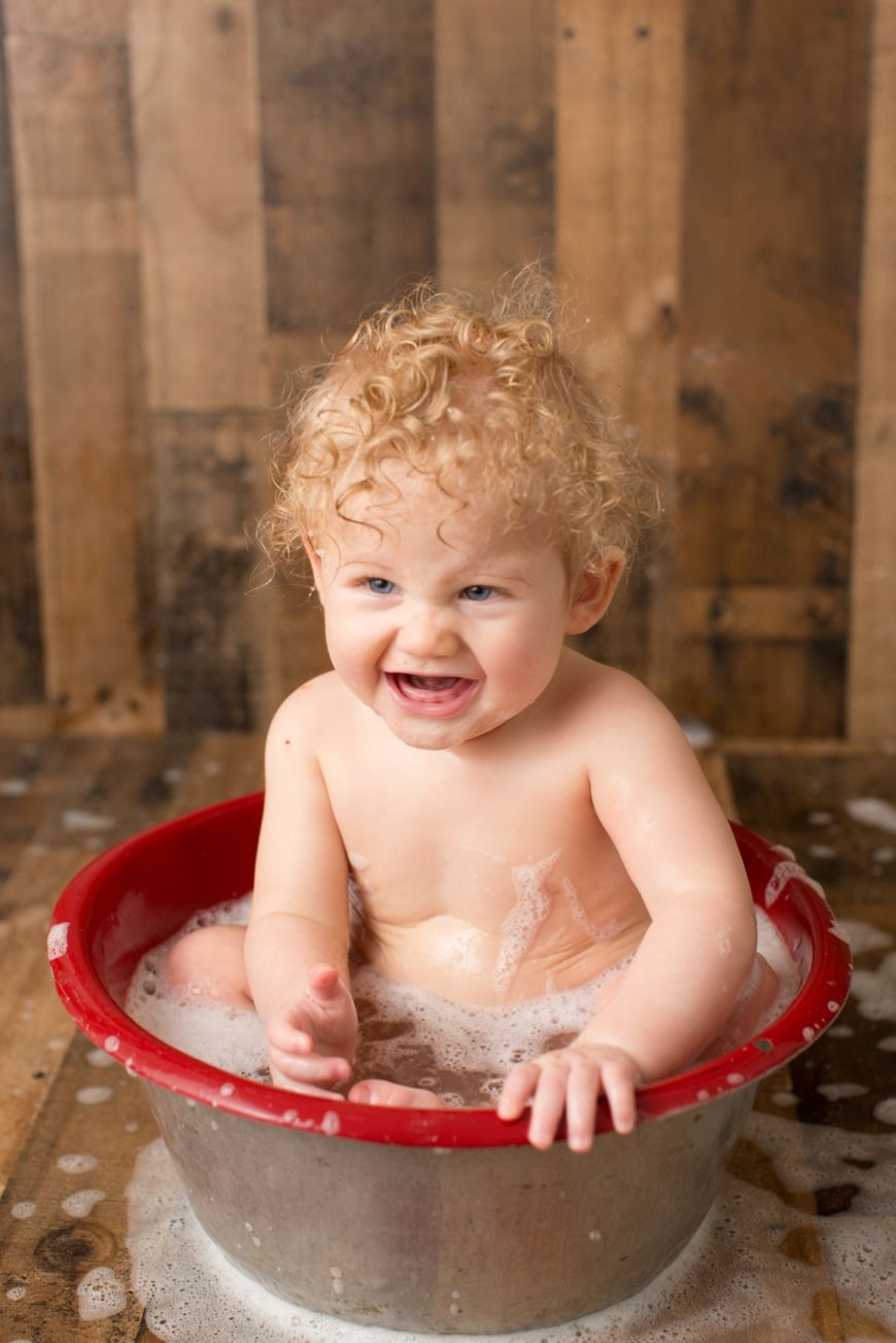 Baby sitting in red-rimmed metal tub with soapy water against wooden backdrop.