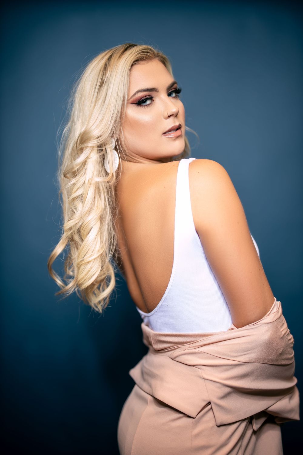 Fashion portrait of a blonde model in white top and beige jacket against blue background.