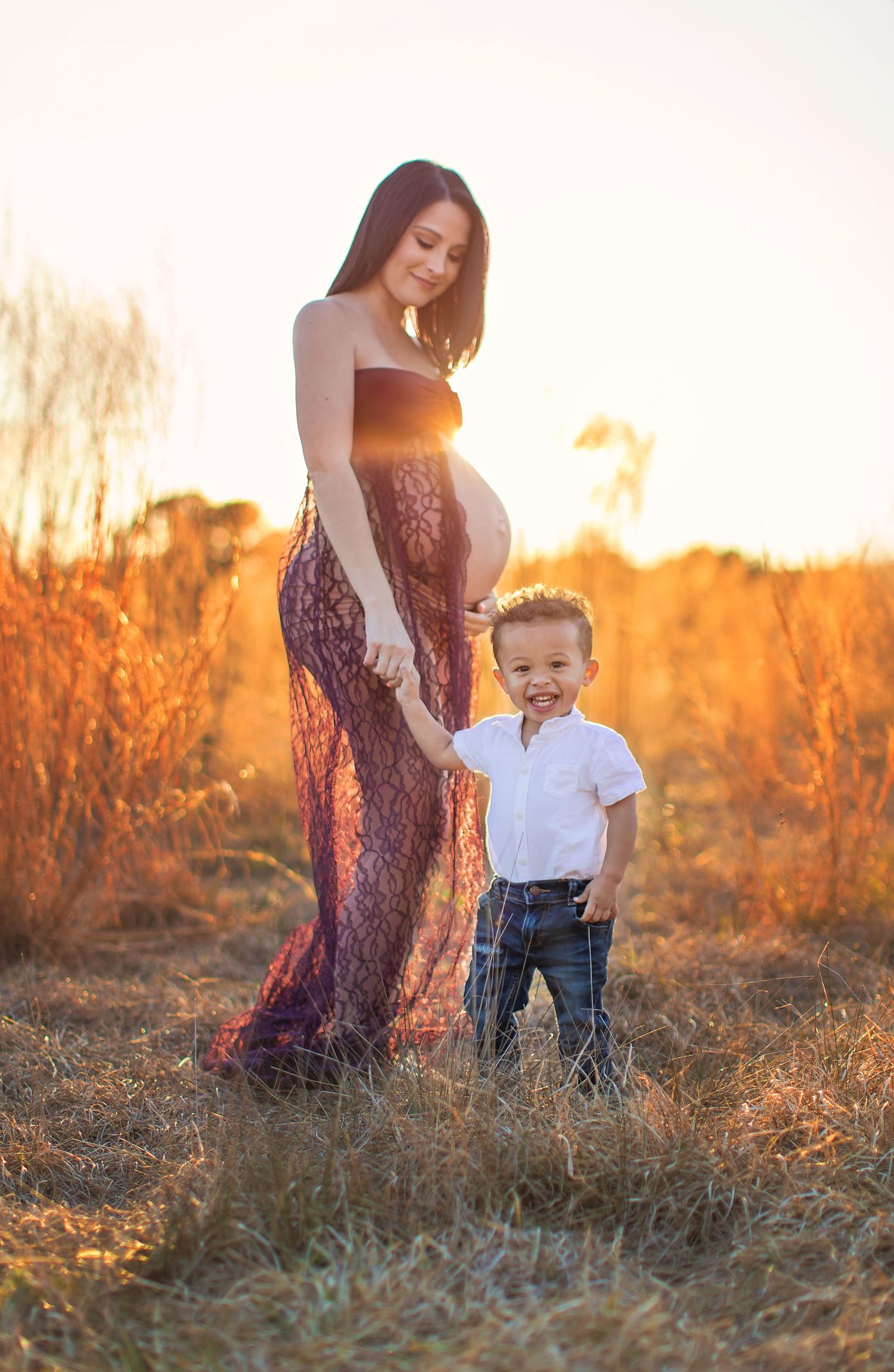 Expectant mother in lace dress poses with young child in golden sunset field.
