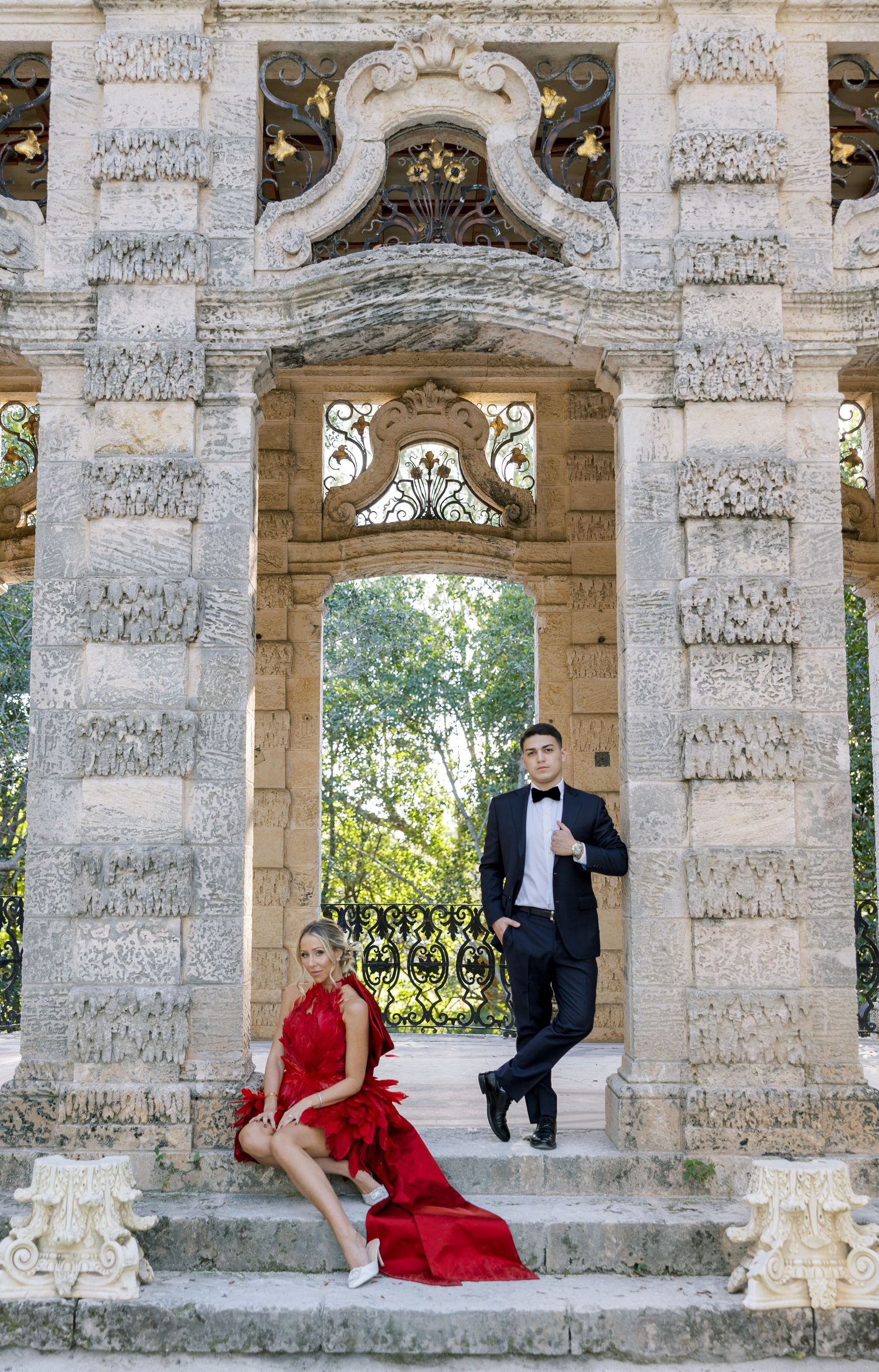 A couple poses elegantly on stone steps of a classical architecture archway wearing formal attire.