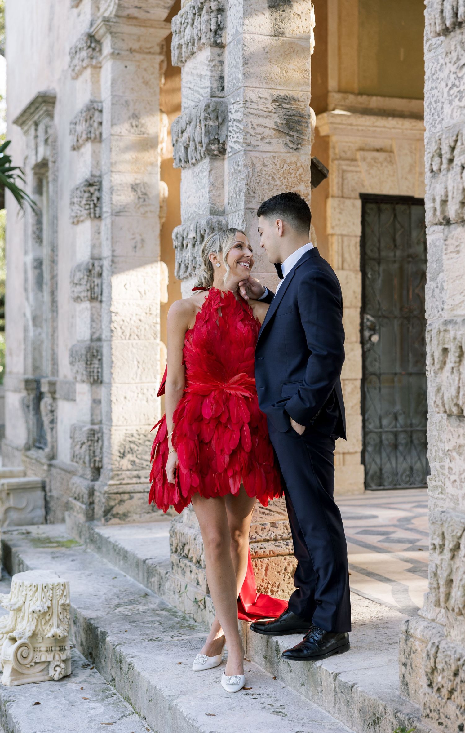 A couple shares a romantic moment between stone pillars, with her wearing a stunning red feathered cocktail dress.