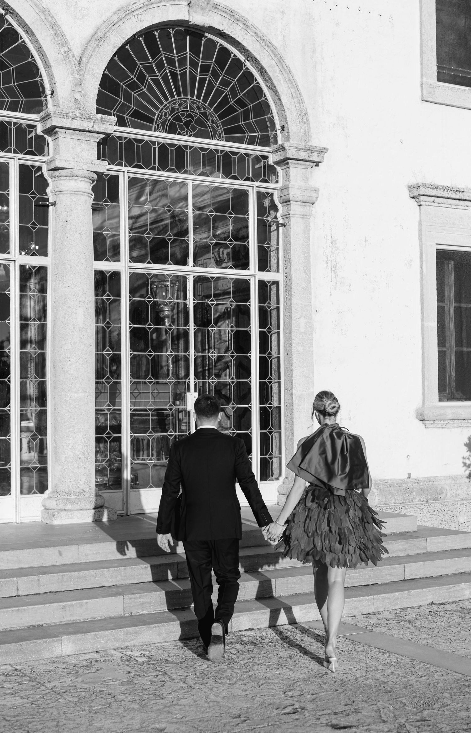Couple walking hand in hand past elegant arched windows in black and white photograph.