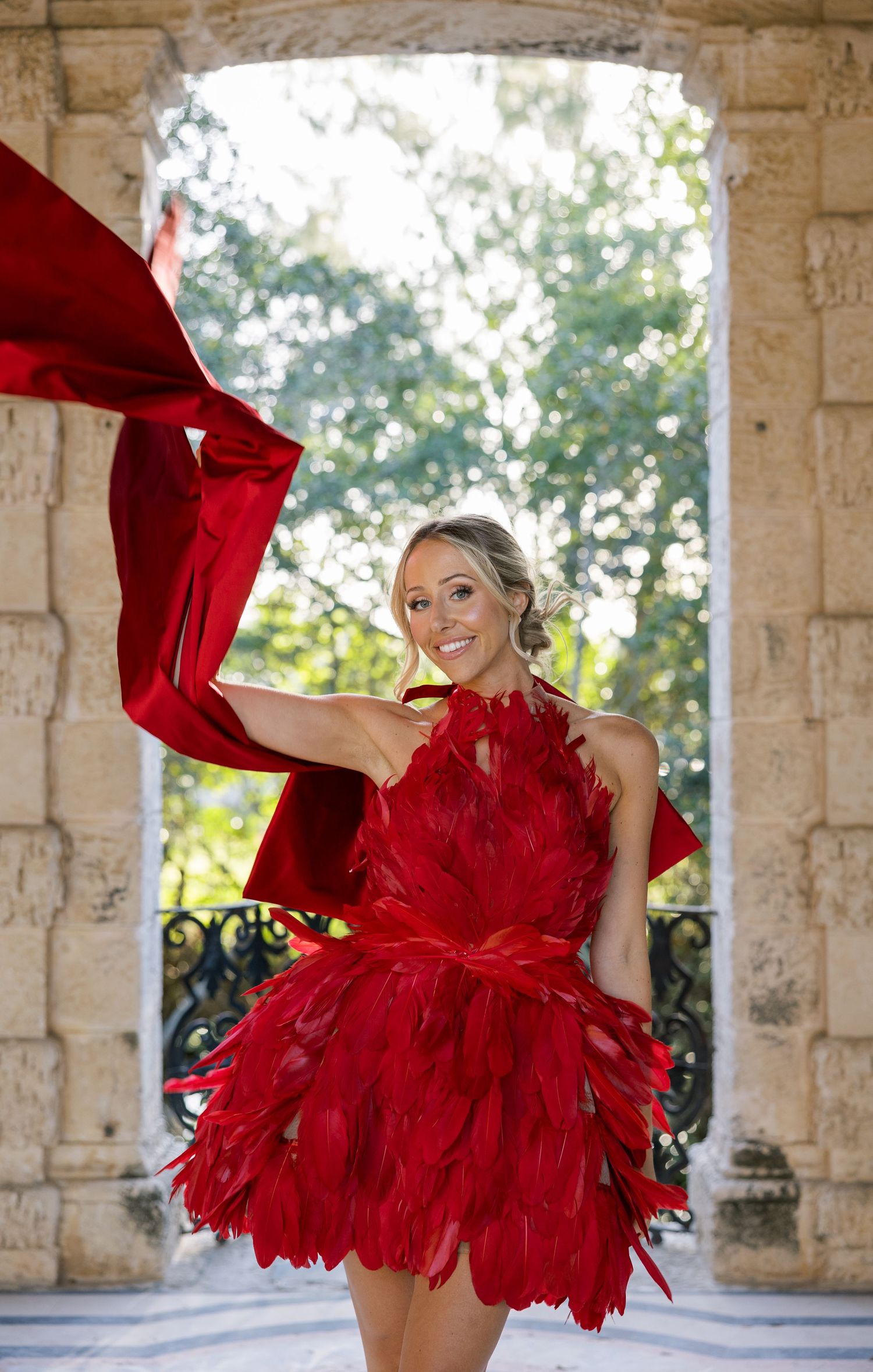 A woman in a red feathered dress twirls dramatically in a stone archway with sunlit gardens behind.