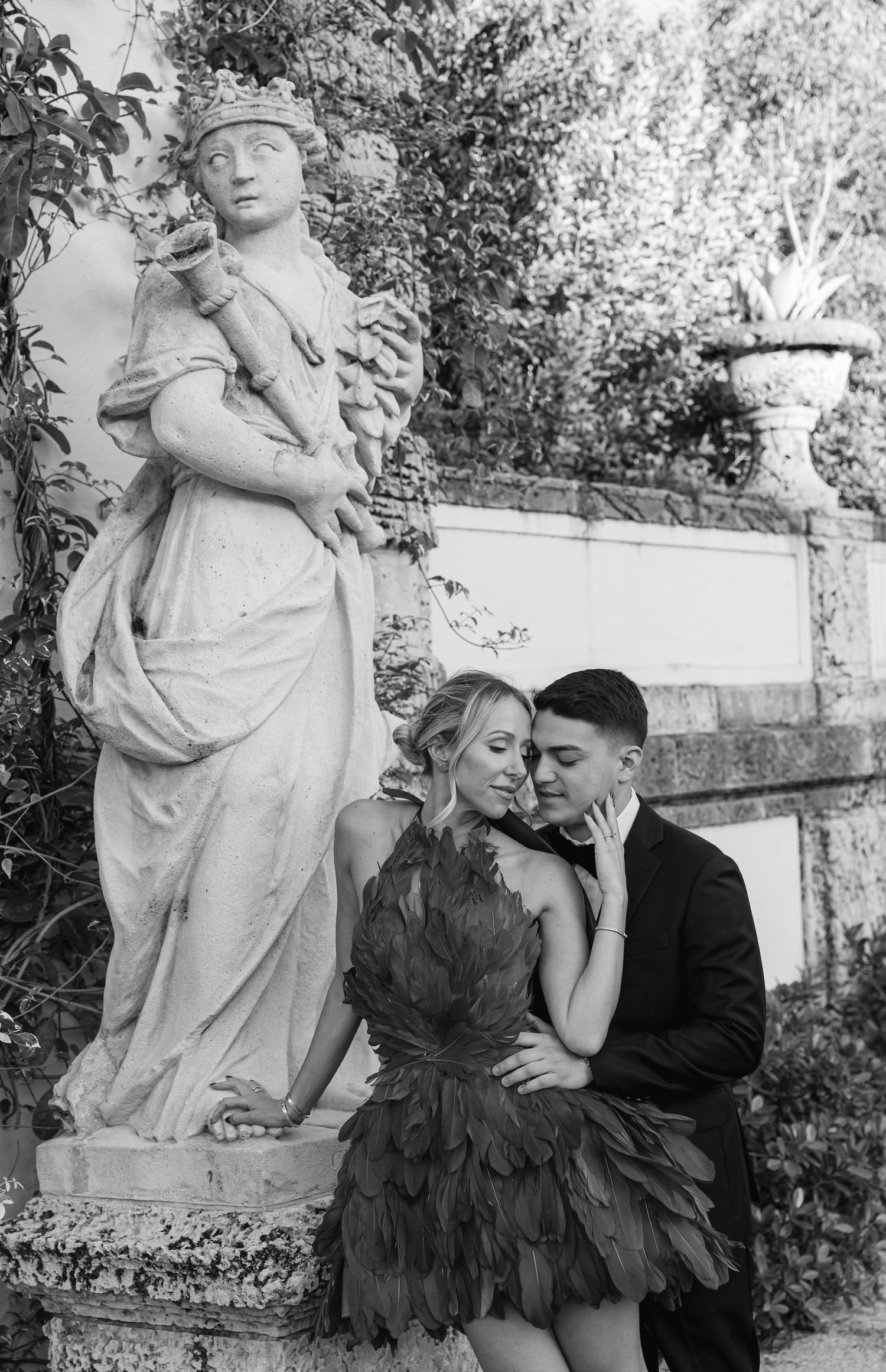 Romantic couple embraces near a classical garden statue in black and white vintage photograph.