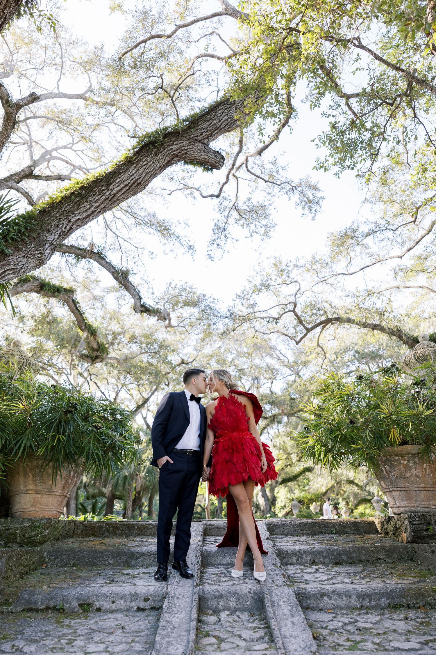 Elegant couple poses under sprawling oak trees on stone path.