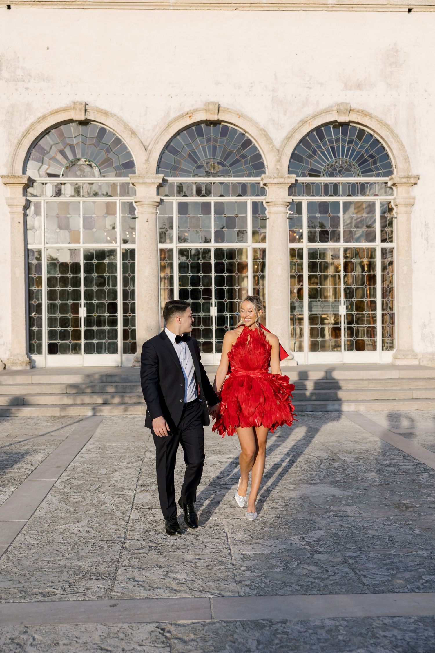 Couple in formal wear strolling in front of classical arched windows.