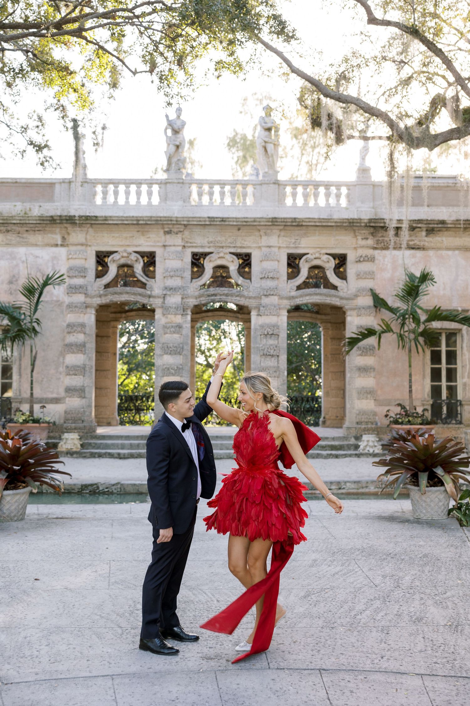 A couple poses playfully by a classical stone facade with tropical gardens in background.