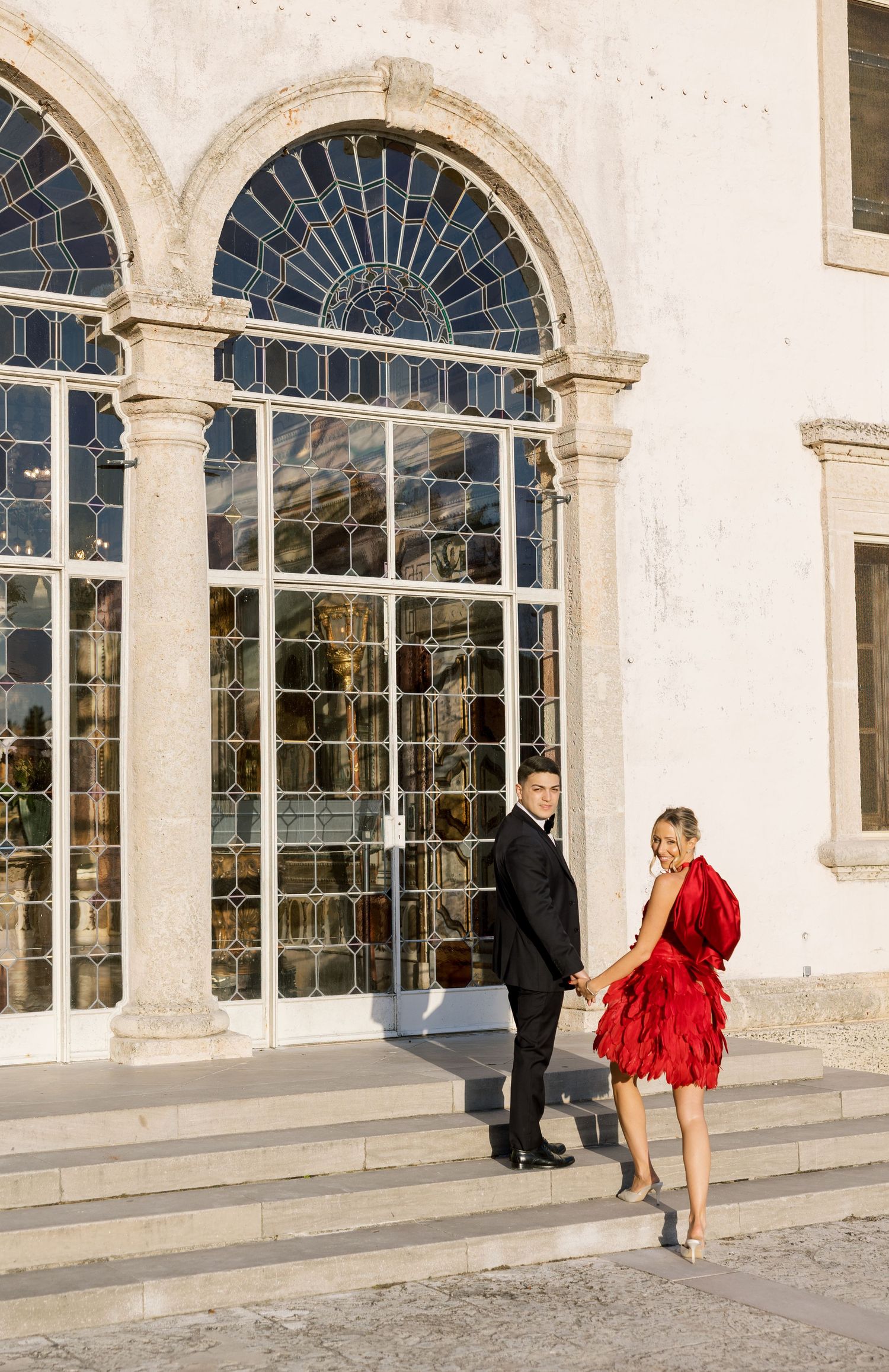 Couple in formal wear poses on steps in front of ornate architectural windows.