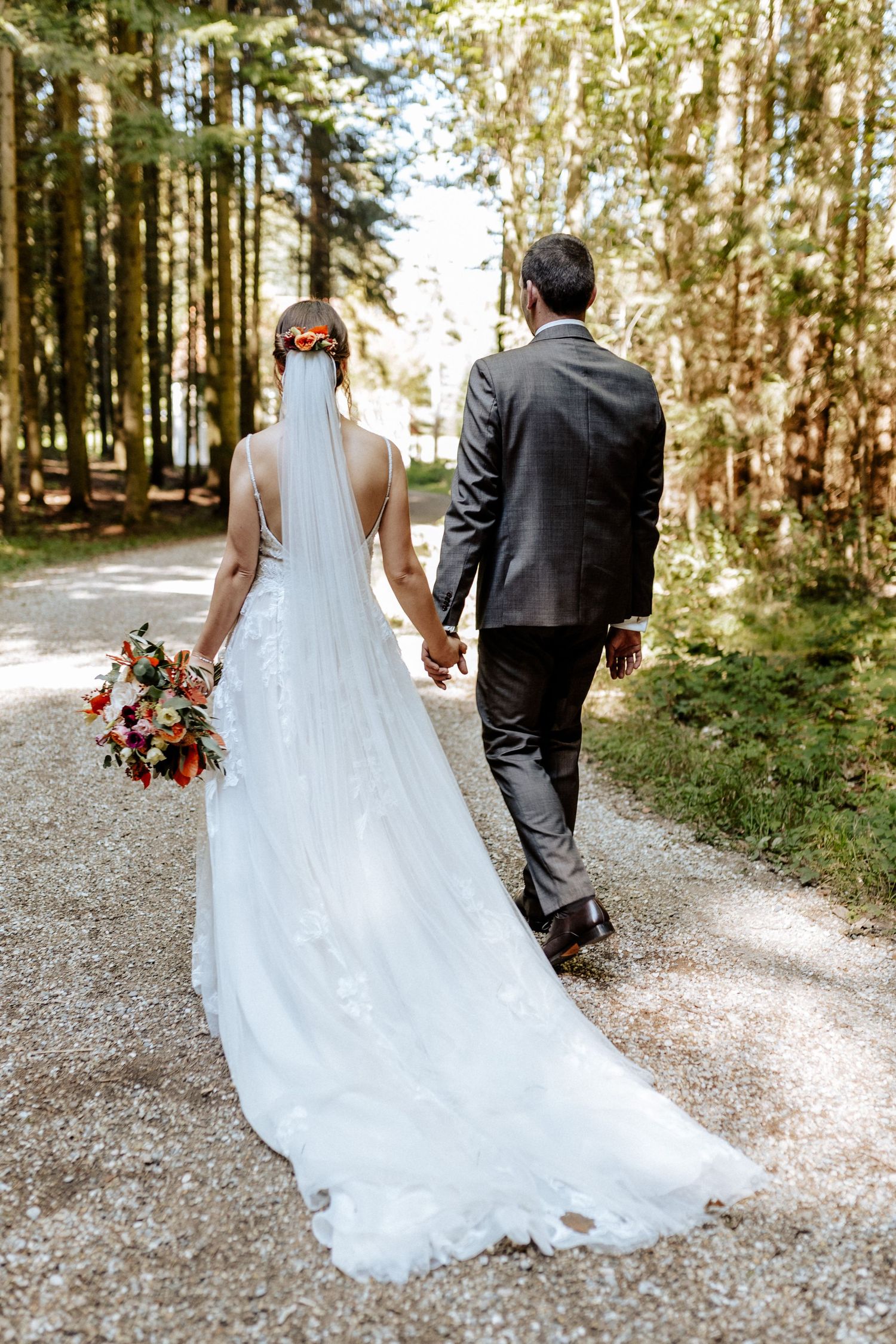 A couple walks down a tree-lined path after their wedding, with the bride carrying a colorful bouquet.