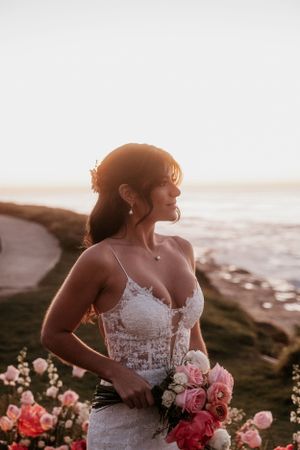 Bride in lace gown holds pink bouquet against coastal sunset backdrop.