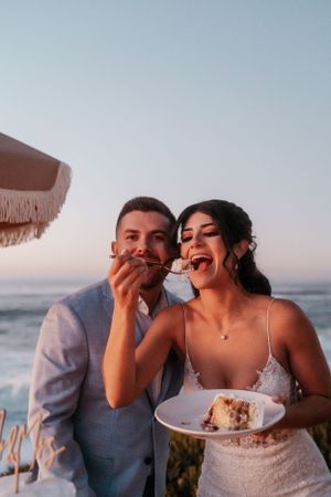 Couple shares wedding cake while laughing at coastal reception.