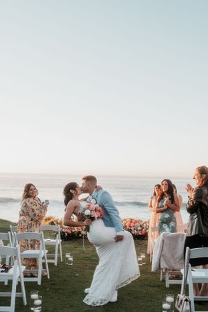 Newlyweds share first dance on oceanfront lawn with wedding guests looking on.