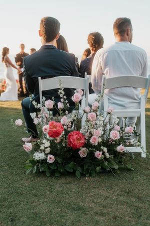 Wedding ceremony seating with pink and white floral arrangements decorating white chairs on a grassy lawn.
