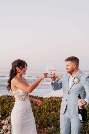 Newlyweds toast with champagne glasses against an ocean backdrop at sunset.