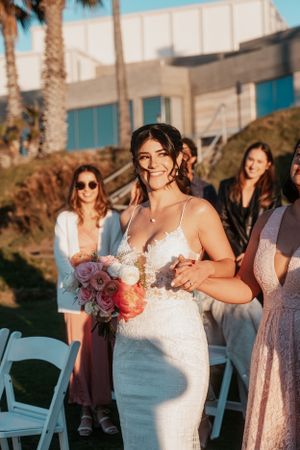 A wedding ceremony participant holds a bouquet of pink flowers while standing among palm trees.