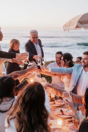 Wedding guests toast at candlelit outdoor reception table by the ocean.