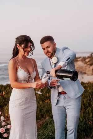 A couple in wedding attire pours champagne at a scenic coastal sunset celebration.