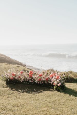 Pink and white floral arrangements line a clifftop wedding ceremony site overlooking the ocean.