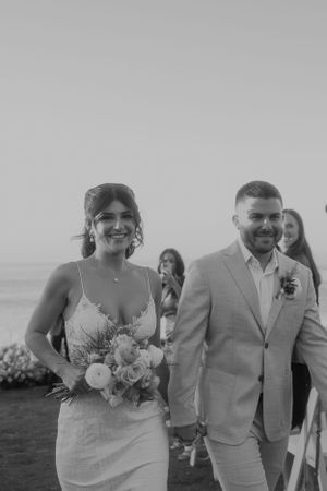 Black and white photo of newlyweds walking together after oceanside ceremony.